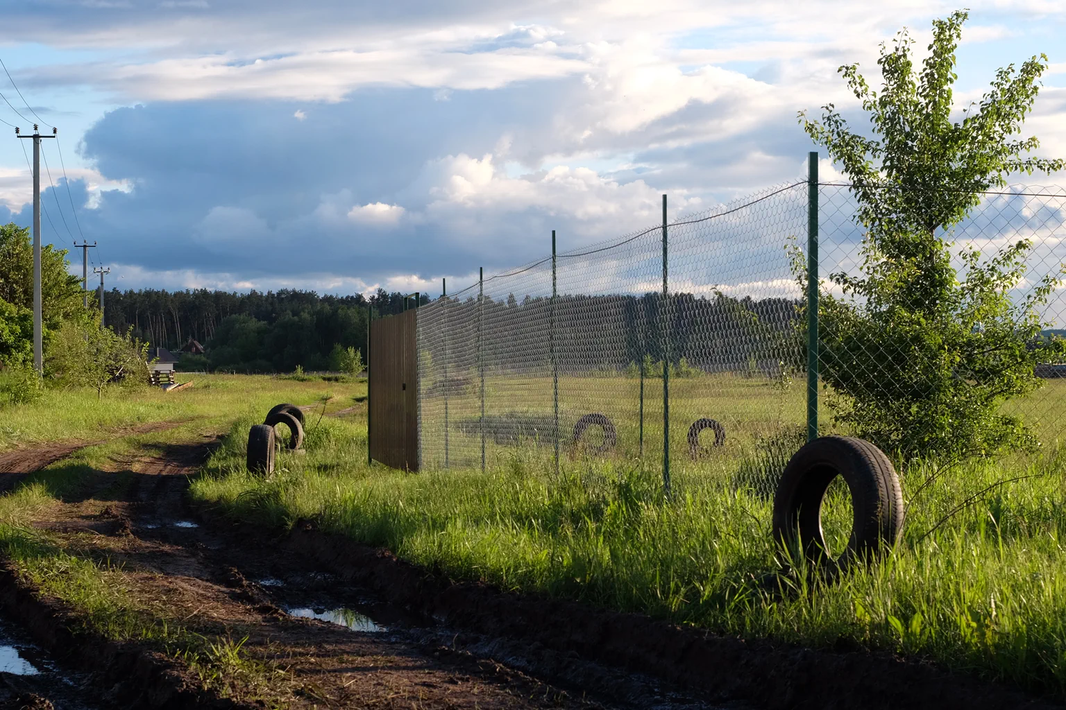 Muddy dirt road alongside a chain-link fence and scattered tires in a rural landscape